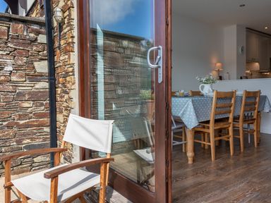 An open glass door leading from a stone patio with a wooden chair to a dining area with a table covered with a polka dot tablecloth and wooden chairs at 7 Island Quay in Salcombe