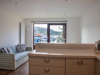A kitchen counter with drawers and a kettle next to a living area with a sofa and pillows by a window showing boats on the water at 7 Island Quay Salcombe