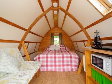 A small wooden room with a bed with red plaid bedding a microwave a sofa and kitchen utensils at Sheep Shed @ Penbugle Organic Farm in St Keyne near Looe