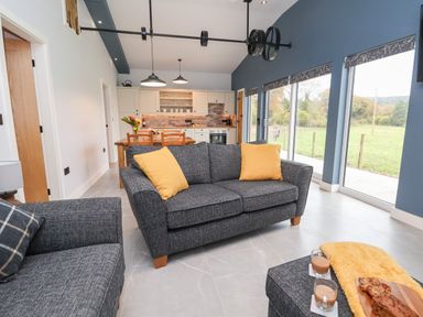 A living area with gray sofas, yellow cushions, a kitchen with wooden dining table, and large windows at Elliw in Bontuchel near Ruthin