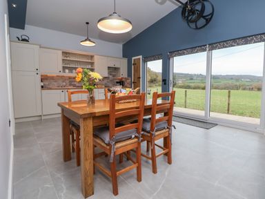 A kitchen with a wooden dining table and chairs set near large windows overlooking a green field at Elliw in Bontuchel near Ruthin