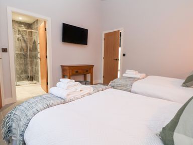 A bedroom with two single beds with white linens and towels on them a wooden table a wall-mounted TV and an open door leading to a bathroom with a shower at Elliw in Bontuchel near Ruthin