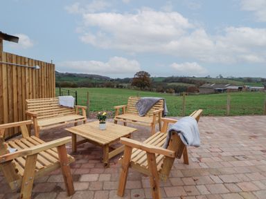 An outdoor patio with wooden benches and chairs around a wooden table with a flower pot overlooking green fields at Elliw in Bontuchel near Ruthin