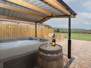 An outdoor covered patio with a hot tub wooden barrel table with a bottle and glasses and wooden seating with a green field in the background at Elliw in Bontuchel near Ruthin