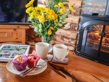A wooden table with two mugs two muffins a magazine yellow flowers and a fireplace in a log cabin at Stag Lodge Holiday Retreat Whitby