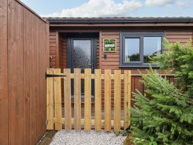 A wooden gate in front of a door and window on a brown wooden building with a welcome sign and greenery nearby at Stag Lodge Holiday Retreat Whitby