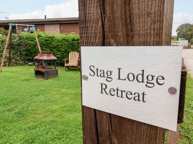 A grassy outdoor area with a fire pit, wooden chair, swing set, and a sign on a wooden post reading Stag Lodge Retreat