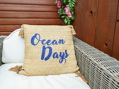 A woven beige cushion with ocean days text on a white sofa with wicker arms and a wooden wall with hanging flowers at Stag Lodge Holiday Retreat in Whitby