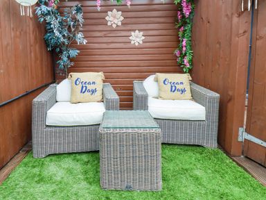 A small outdoor seating area with two wicker chairs with white cushions and ocean days pillows with a wicker table between them at Stag Lodge Holiday Retreat in Whitby
