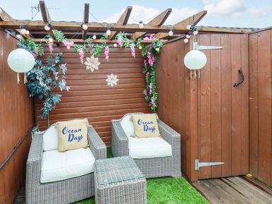An outdoor seating area with two wicker armchairs and a matching table under a wooden pergola decorated with hanging lights and flowers at Stag Lodge Holiday Retreat in Whitby
