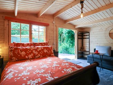 A bedroom with floral bedding a window a sofa with a side table and an open door showing greenery at Log Cabin - Amber Glow in Glastonbury
