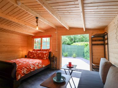 A wooden interior room with a bed with red floral bedding a small side table a gray sofa two small tables with cups and an open doorway showing a fenced garden at Log Cabin - Amber Glow in Glastonbury