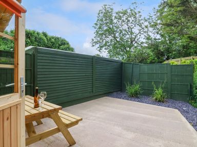 A small outdoor patio area with wooden picnic table glass wind and bottle surrounded by green painted wooden fences and plants at Log Cabin - Amber Glow in Glastonbury
