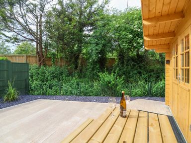 An outdoor patio area with a wooden picnic table holding a bottle and two wine glasses next to a wooden cabin surrounded by greenery at Log Cabin - Amber Glow in Glastonbury