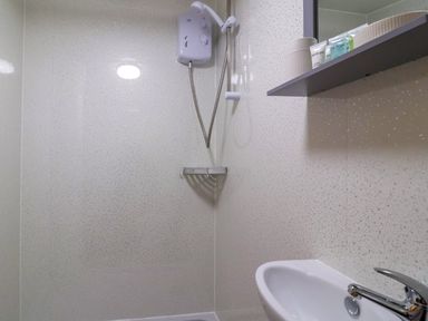 A bathroom shower area with an electric shower unit a small white sink and a shelf with toiletries at Log Cabin - Amber Glow in Glastonbury