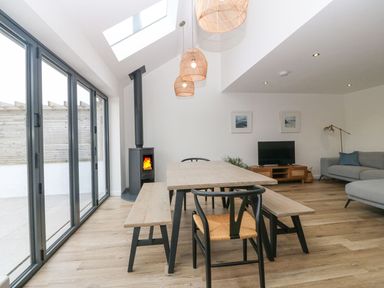 A dining area with a wooden table and benches next to a living room with a gray sofa and a TV at 11 Capel Farm in Trearddur Bay