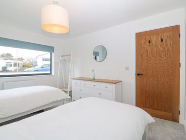 A bedroom with two single beds a wooden door a white chest of drawers a round mirror and a window at 11 Capel Farm in Trearddur Bay