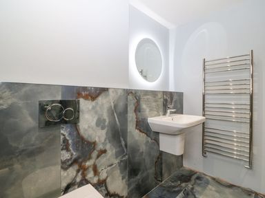 A bathroom with a wall-mounted sink a circular illuminated mirror a heated towel rail and dark marble tiles at 11 Capel Farm in Trearddur Bay