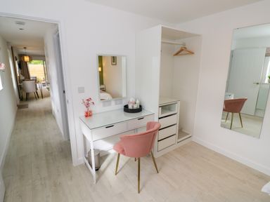 A white vanity desk with a mirror and pink chair next to an open wardrobe and a wall mirror at The Croft in Shipston-On-Stour