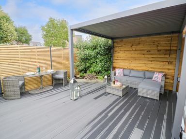 An outdoor patio with wicker furniture including a table with two chairs and a sectional sofa under a pergola at The Croft in Shipston-On-Stour