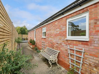 A narrow outdoor pathway with a wooden bench and potted plants along a brick wall at The Croft in Shipston-On-Stour