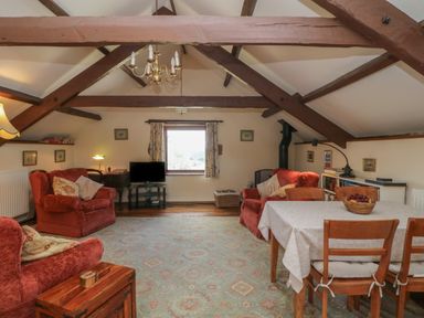 An attic living room with exposed wooden beams two red armchairs a dining table with fruit and a television at Beckaford Cottage in Bovey Tracey