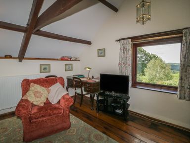 A room with a red armchair a wooden desk with a lamp a chair a television on a glass stand and a window showing trees at Beckaford Cottage in Bovey Tracey