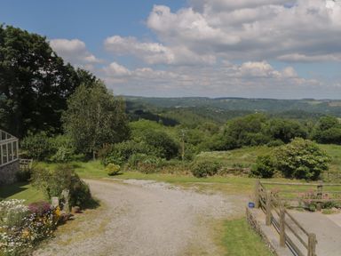 An outdoor scene with a gravel path flowers trees and a wooden fence with hills and clouds in the background at Beckaford Cottage in Bovey Tracey