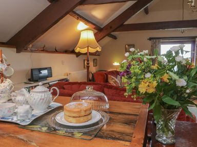 A living room with a wooden table holding a cake tea set and a vase of flowers at Beckaford Cottage in Bovey Tracey