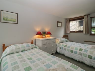 A bedroom with two single beds a white dresser with lamps and a window showing an outside stone wall at Beckaford Cottage in Bovey Tracey