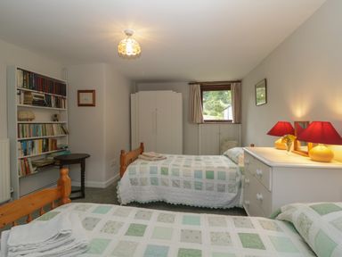 A bedroom with two single beds, a white dresser with red lamps, a bookshelf, and a window at Beckaford Cottage in Bovey Tracey