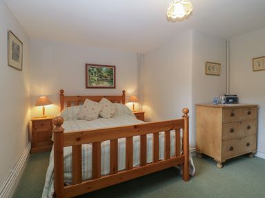 A bedroom with a wooden bed frame two bedside tables with lamps a wooden dresser and framed pictures on the walls at Beckaford Cottage in Bovey Tracey