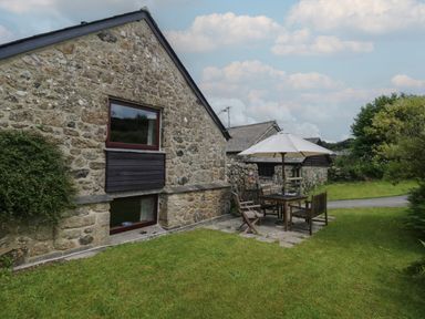 A stone exterior wall of a cottage with wooden window frames and outdoor furniture with an umbrella on a grass lawn at Beckaford Cottage in Bovey Tracey