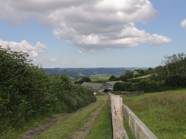A country lane with wooden fence and bushes leading to farm buildings with hills and clouds in the background at Beckaford Cottage Bovey Tracey