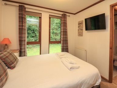 A bedroom with a double bed with white sheets and plaid pillows two folded towels on the bed a wall-mounted television two windows with plaid curtains and a bedside table with a lamp at Templar Rise 16 - Devon Comfort Finlakes Holiday Park near Chudleigh