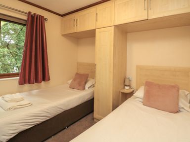 A bedroom with two single beds a window with red curtains and overhead wooden cabinets at Templar Rise 16 - Devon Comfort Finlakes Holiday Park near Chudleigh