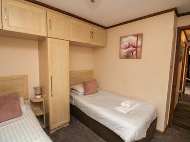 A bedroom with two single beds, overhead cabinets, a bedside table with a lamp, a flower painting on the wall at Templar Rise 16 - Devon Comfort Finlakes Holiday Park near Chudleigh