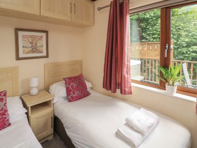 A bedroom with two single beds a bedside table with a lamp a window with red curtains and a potted plant on the windowsill at Templar Rise 16 - Devon Comfort Finlakes Holiday Park near Chudleigh