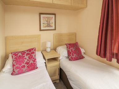 A bedroom with two single beds a bedside table with a lamp a framed picture on the wall and red curtains at Templar Rise 16 - Devon Comfort Finlakes Holiday Park near Chudleigh