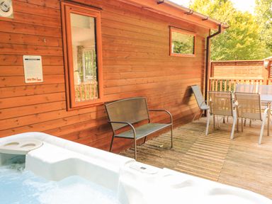 An outdoor deck with a hot tub bench a metal chair a dining table with chairs next to a wooden cabin at Deer Park 9 - Copse Corner Finlakes Holiday Park near Chudleigh