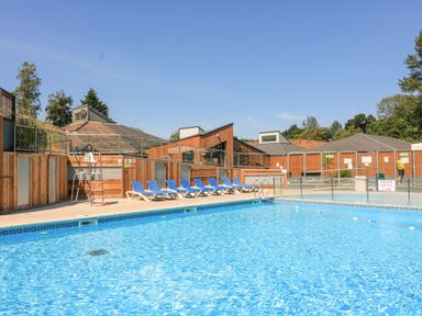 An outdoor swimming pool with blue lounge chairs and wooden buildings in the background at Deer Park 9 - Copse Corner Finlakes Holiday Park near Chudleigh