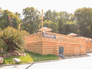 An outdoor wooden fenced area with umbrellas and a sign for The Hub Shop surrounded by trees at Finlakes Holiday Park near Chudleigh