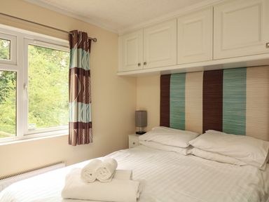 A bedroom with a bed, folded towels on the bed, a window with patterned curtains, bedside table with a lamp and overhead storage cupboards at The Brambles 17 - Conkers Lodge Finlakes Holiday Park near Chudleigh
