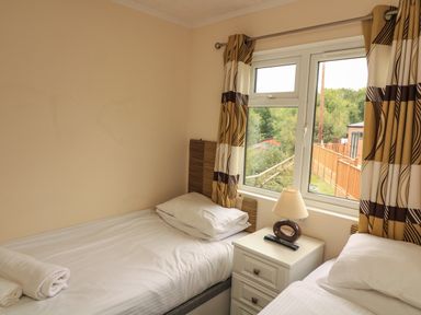 A small bedroom with two single beds separated by a white nightstand with a lamp and remote near a window with patterned curtains at The Brambles 17 - Conkers Lodge Finlakes Holiday Park near Chudleigh