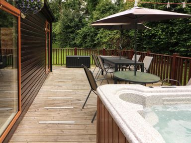 An outdoor wooden deck with a hot tub dining table chairs umbrellas and railing at The Brambles 17 - Conkers Lodge Finlakes Holiday Park near Chudleigh
