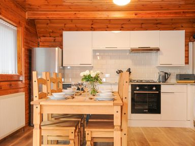 A kitchen with a wooden dining table set with dishes and glasses surrounded by wooden chairs and white cabinets with appliances at Lodge 32 - Cowslip Finlakes Holiday Park near Chudleigh