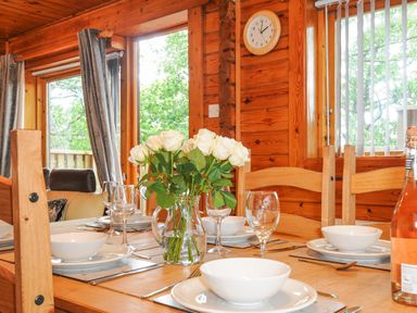 A dining table set with white bowls and wine glasses with a vase of white roses on the table in a wooden interior dining room at Lodge 32 - Cowslip Finlakes Holiday Park near Chudleigh