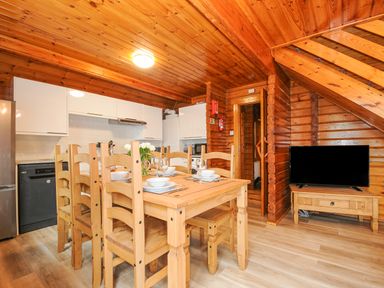 A dining area with wooden table and chairs adjacent to a kitchen with white cabinets and a television on a wooden stand under stairs at Lodge 32 - Cowslip Finlakes Holiday Park near Chudleigh