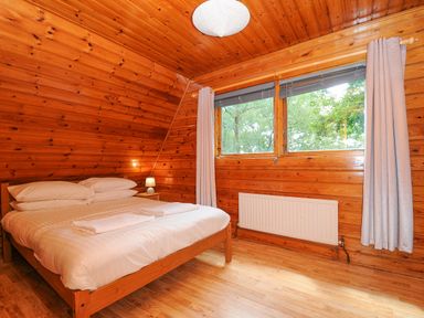 A bedroom with a double bed next to a window with curtains and a small bedside table with a lamp in a wooden interior at Lodge 32 - Cowslip Finlakes Holiday Park near Chudleigh