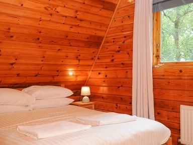 A bedroom with a bed with white linens two folded towels bedside table lamp wooden walls and a window with curtains at Lodge 32 - Cowslip Finlakes Holiday Park near Chudleigh